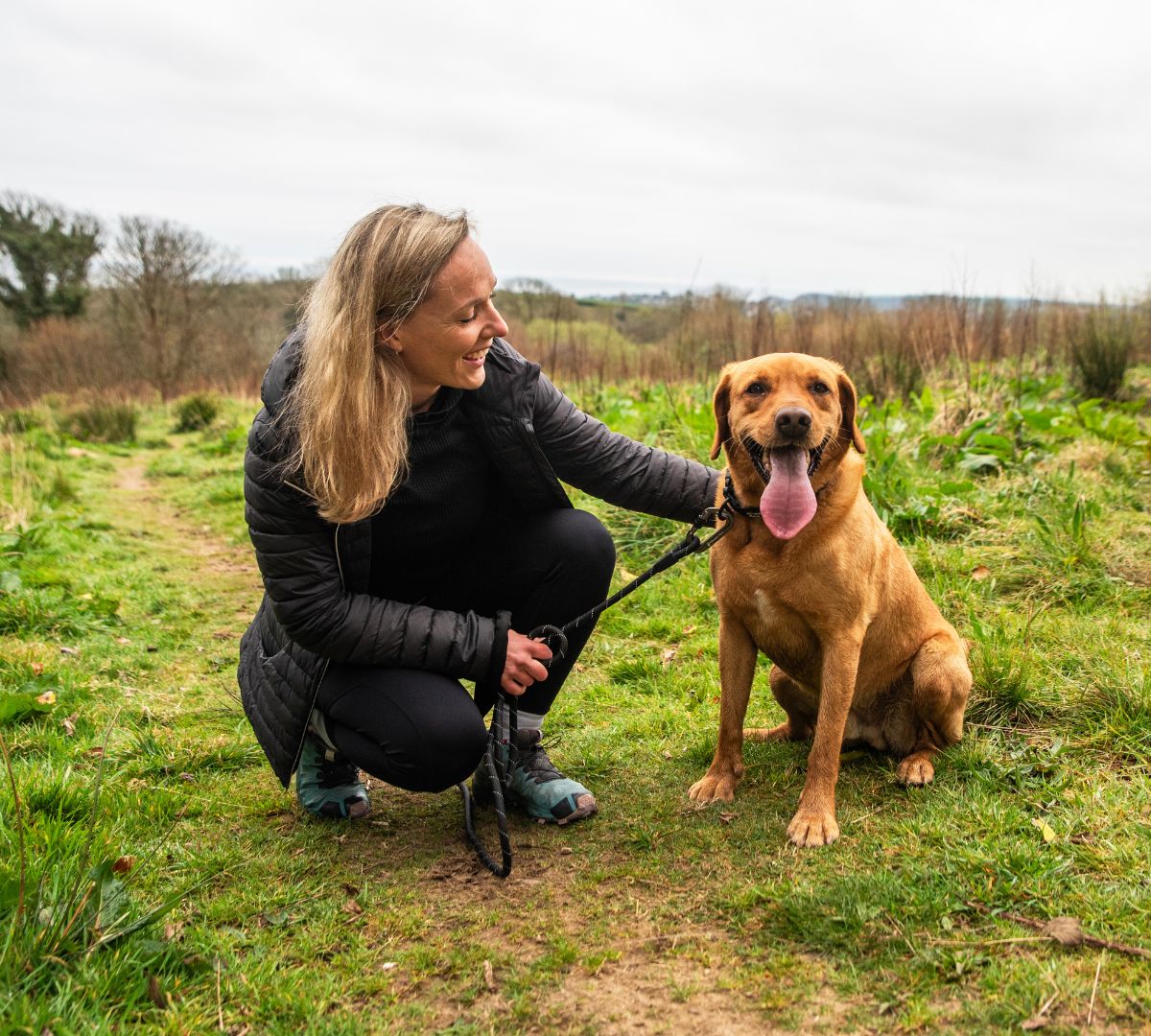 Woman kneeling next to a smiling Labrador on a countryside trail, both enjoying a break during a walk with a Mountain Paws lead.