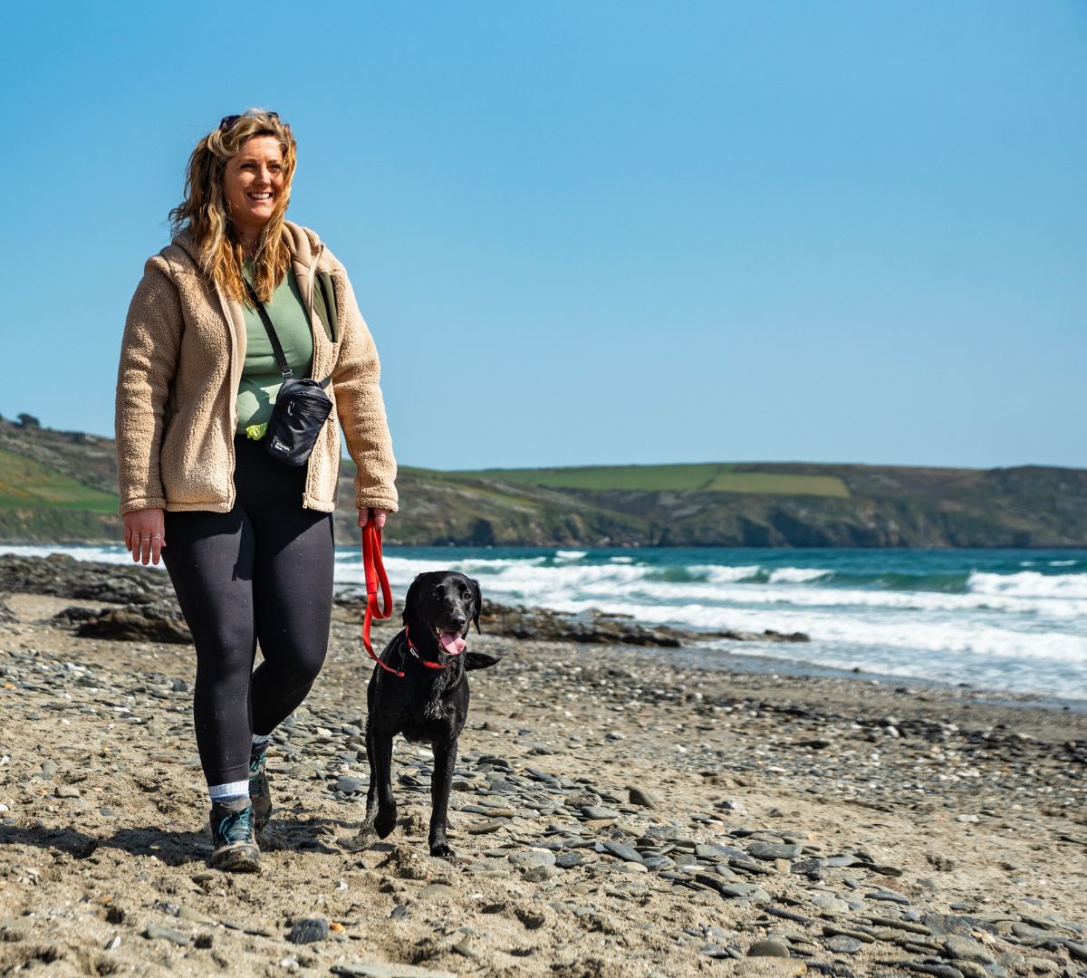 Smiling woman walking a black dog along a scenic beach, using a red Mountain Paws lead.